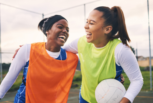 Two girl teammates who play netbal embrace into a hug while smiling and laughing with each other